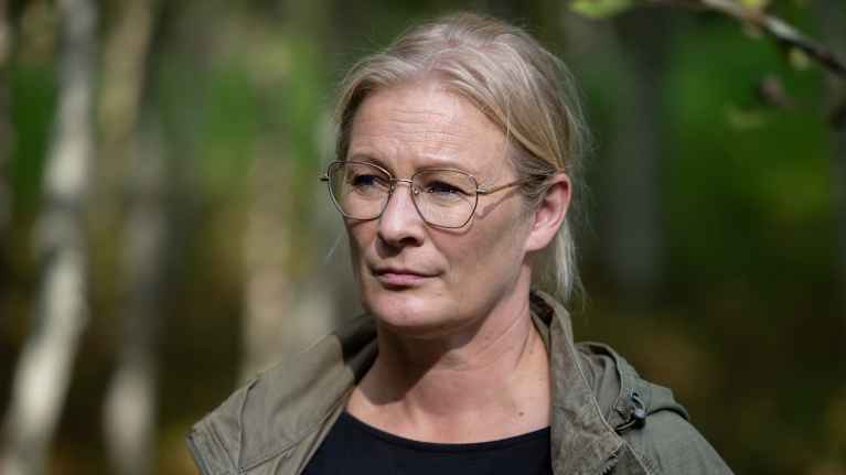 En kvinna med ljust bakåtkammat hår och glasögon iklädd en grön jacka står i en skog. Bakgrunden är suddig, med synliga träd och grönska.