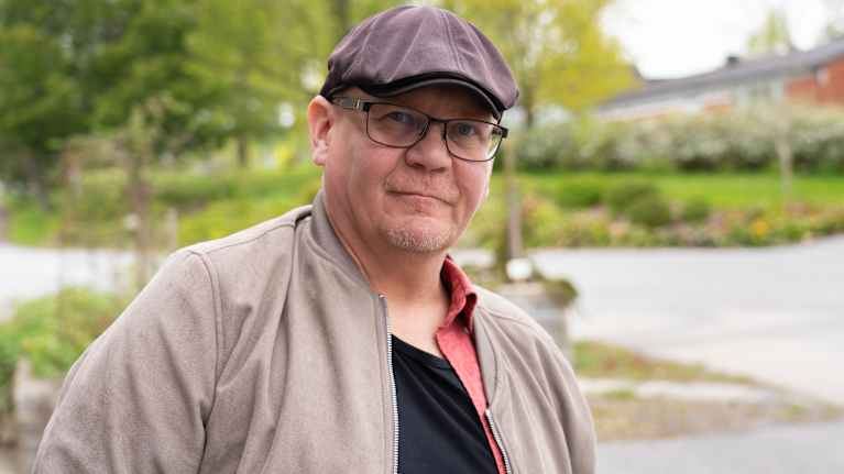 Medelålders man utomhus, tittar in i kameran med ett snett leende. Mannen har glasögon, beige uppknäppt jacka och keps.