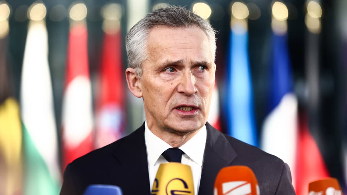 Jens Stoltenberg i mörk kostym står och talar med journalister. I förgrunden syns mikrofoner. Han tittar åt sidan.