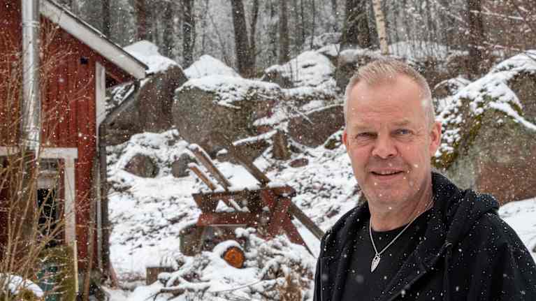 En man står framför en röd stuga i snöoväder.