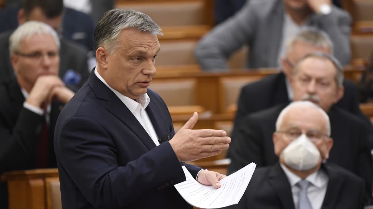 Viktor Orban puhuu paperit kädessään. Taustalla mies hengityssuojuksessa.