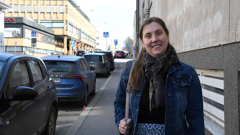 En kvinna med handväska står på en gata i en stad och ser glad ut.