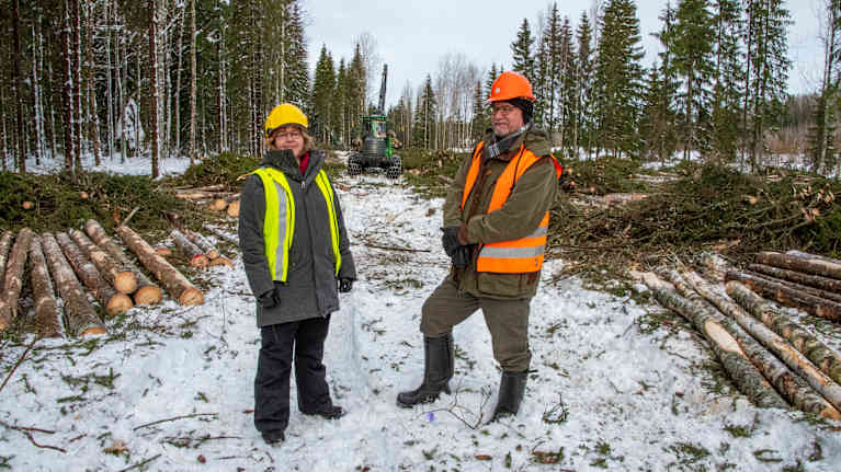 Saija Huuskonen och Markku Rantala står framför en skogsmaskin som håller på att avverka skog.