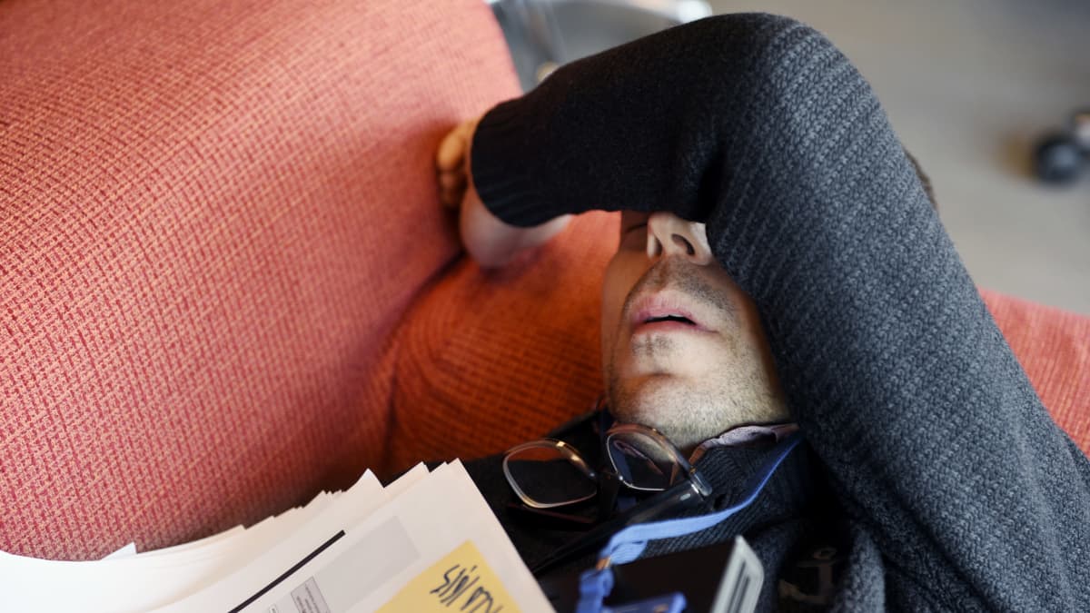 A man with a stubbly beard lies on a couch with his glasses and work papers spread out and an arm across his eyes.