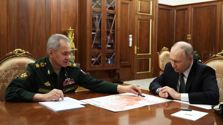 Vihreässä univormussa oleva Sergei Šoigu esittelee mustassa puvussa olevalle Vladimir Putinille karttaa pöydän ääressä. .