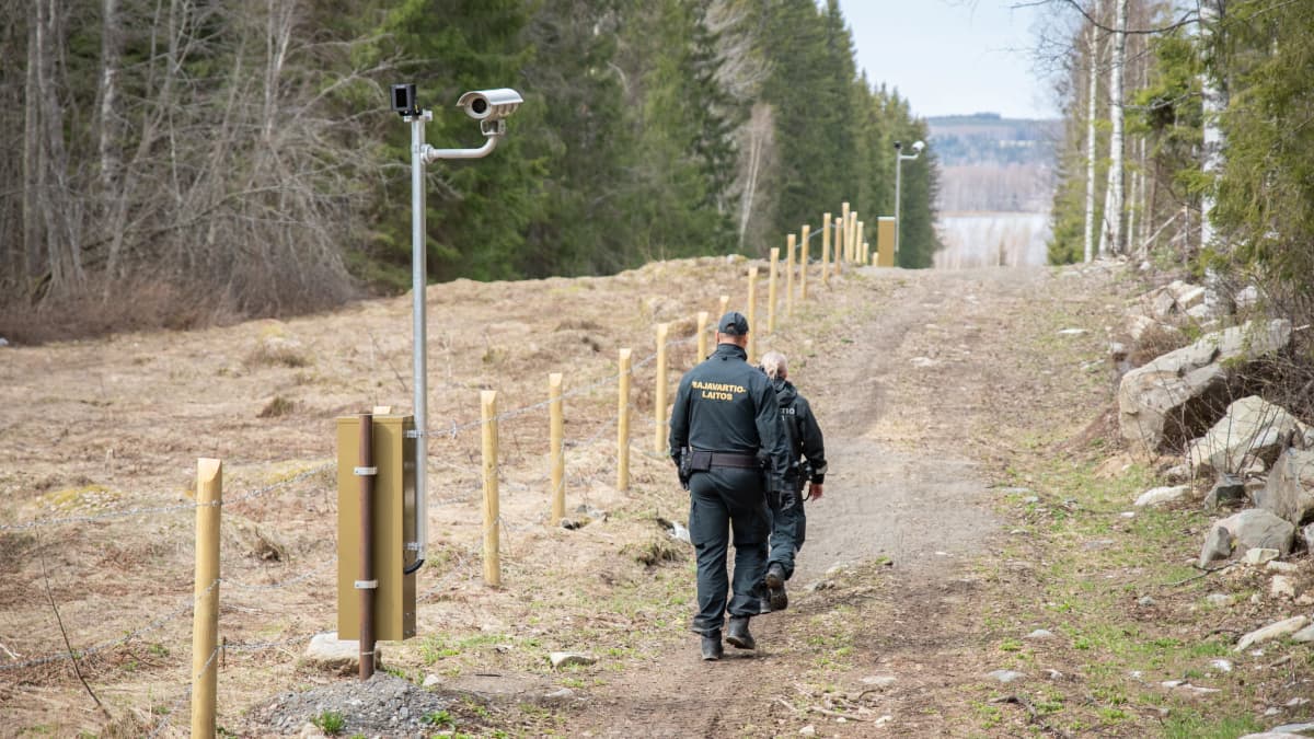 Kaksi rajavartijaa kävelee raja-aukolla piikkilanka-aidan ja valvontakameratolpan vieressä.