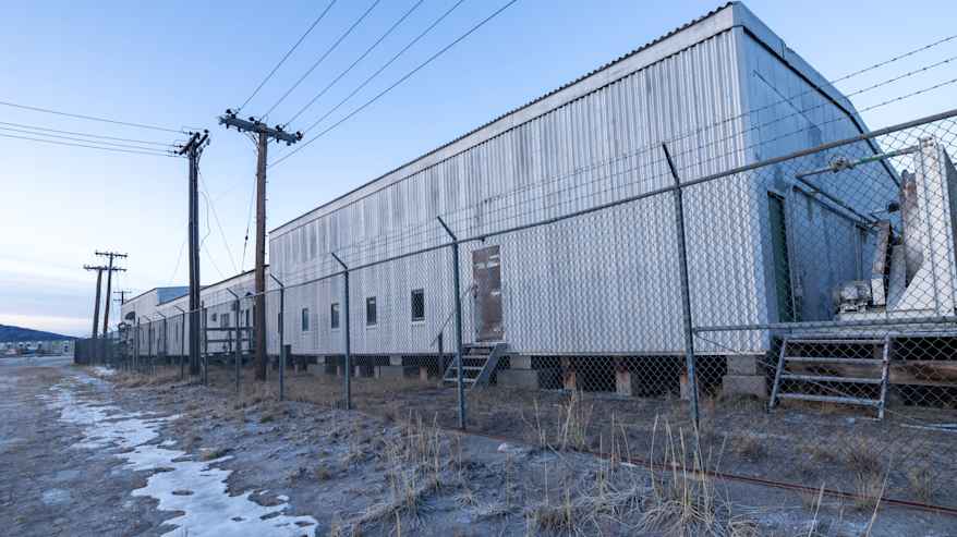 Teräslangalla ⁢aidattu ja piikkilangalla suojattu vanha⁣ peltinen pitkä rakennus. 