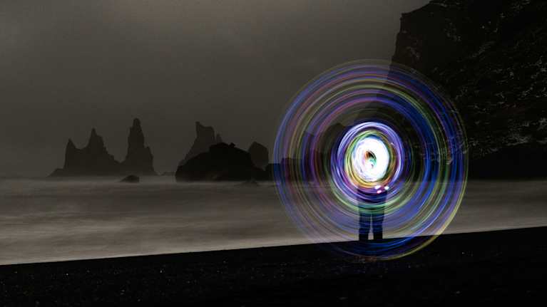 En person står på en mörk strand och snurrar en lysande färgglad cirkel, vilket skapar ett hypnotiskt ljusmönster. 