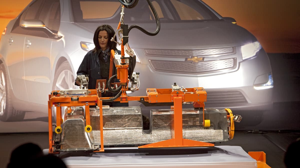 Pitkähiuksinen nainen valmistaa koneella akkua työhaalarissa ja käyttää jonkinlaista poraa. Taustalla näkyy General Motorsin Chevrolet Volt -sähköauto.