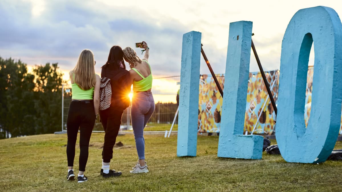 Kolme nuorta naista Ilosaarirock-kirjaimien vierellä selfietä ottamassa. Aurinko paistaa.