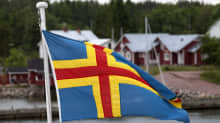 En åländsk flagga i förgrunden, röda hus med vita knutar i bakgrunden.