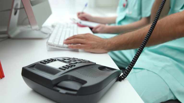 A healthcare professional is talking on the phone. The picture shows a black landline phone on a table and the employee’s hands on the keyboard.