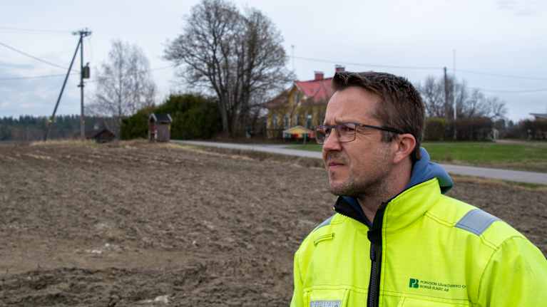 En man i gul jacka står framför en väg med ett gult hus intill en åker.