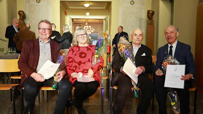 Kjell Nybacka, Pia Lahdenmäki, Joel Lenlund ja Henrik Fågelberg istuvat juhlallisesti tilaisuudessa, pidellen kunniakirjoja ja kukkakimppuja. 