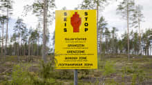 A yellow sign in a thinly forested area, showing a red hand and the words SEIS / STOP plus BORDER ZONE and other text in 5 languages.