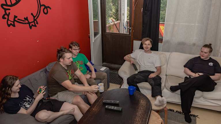 Five young men in T-shirts relaxing on two sofas with a cartoon pig on a wall and a window and balcony in the background.