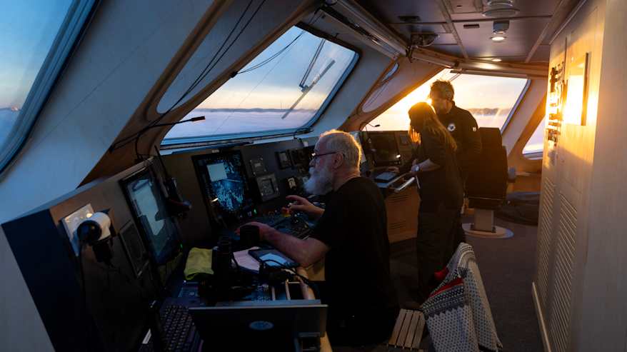 Yksi ihminen työskentelee aluksen yläkerroksessa laitteiston edustalla, kaksi ihmistä keskustelee taustalla.