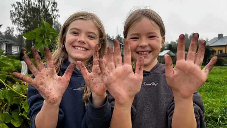 Två skolelever ler framför kameran och visar sina jordiga händer. De står vid skolträdgården vid Kvevlax lärcenter.