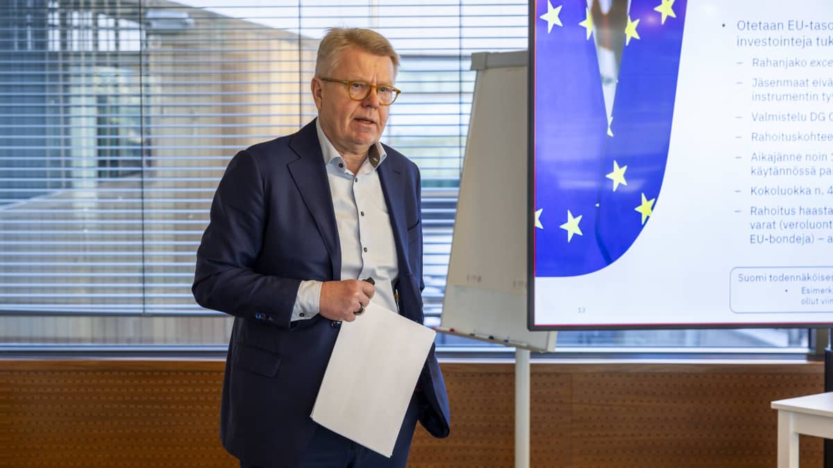 A middle-aged man with light brown hair and glasses and a blue suit with an open-neck shirt is holding paper in a seminar room.