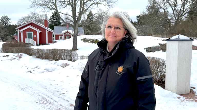 En kvinna står utomhus med snö omkring sig. I bakgrunden röda hus, träd och buskar. 