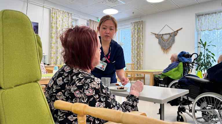 A young Asian woman in a blue staff uniform serves an older seated woman, seen from behind, with two other elderly people in the background.