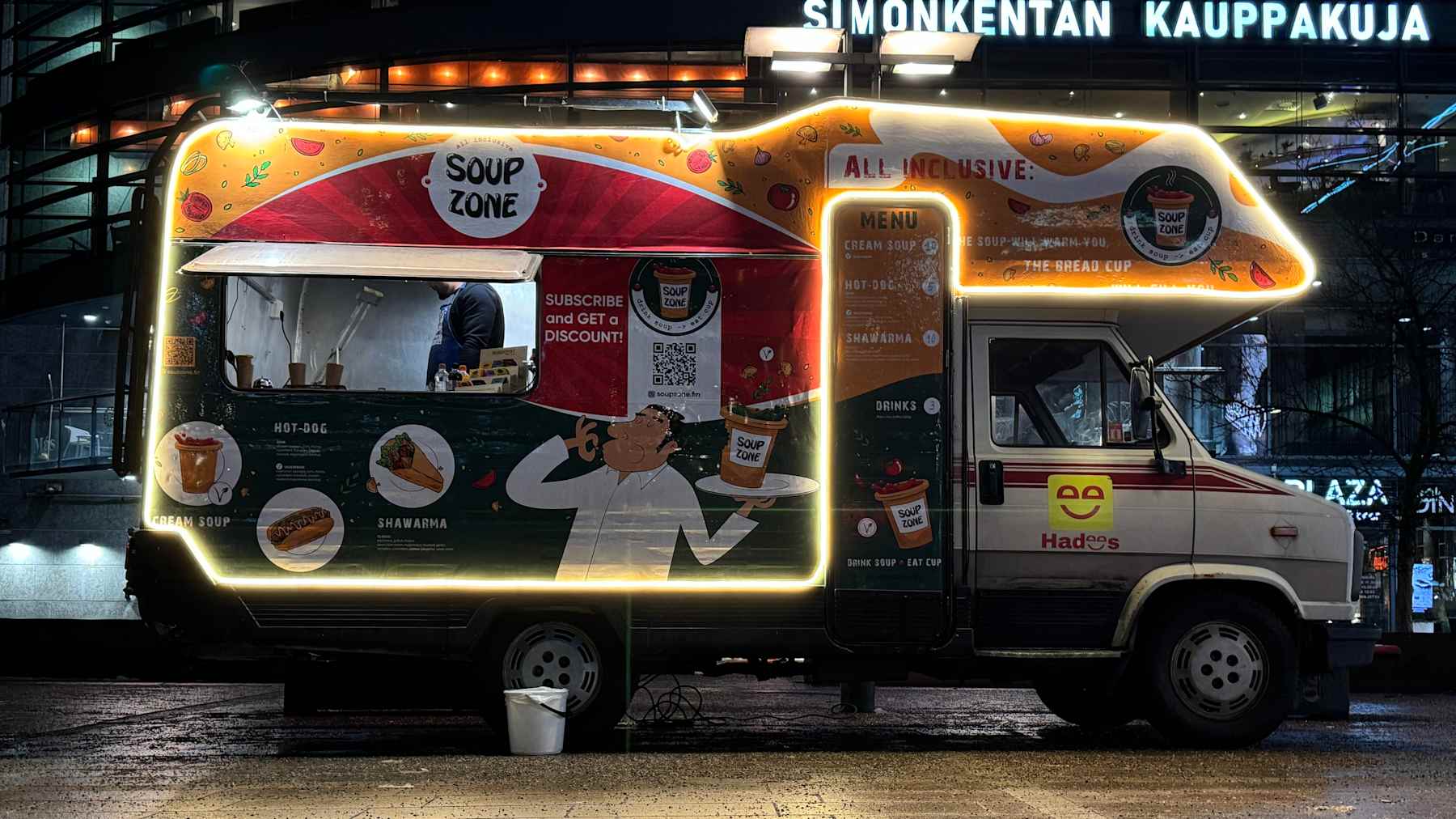a minivan is parked in the square, a light is on in the window and a man is preparing food.