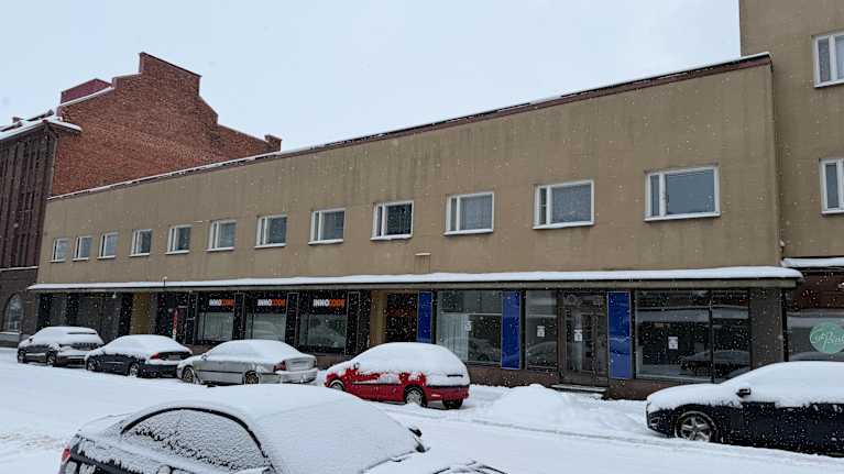 Rakennus taustalla ja lumisia autoja tienvarressa rakennuksen edustalla.