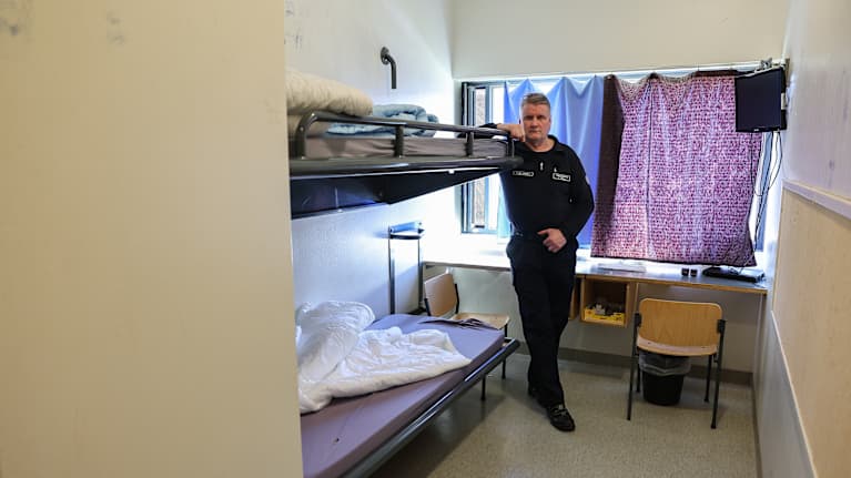 A man in a dark uniform stood in a small room with two bunk beds, a table and a TV.