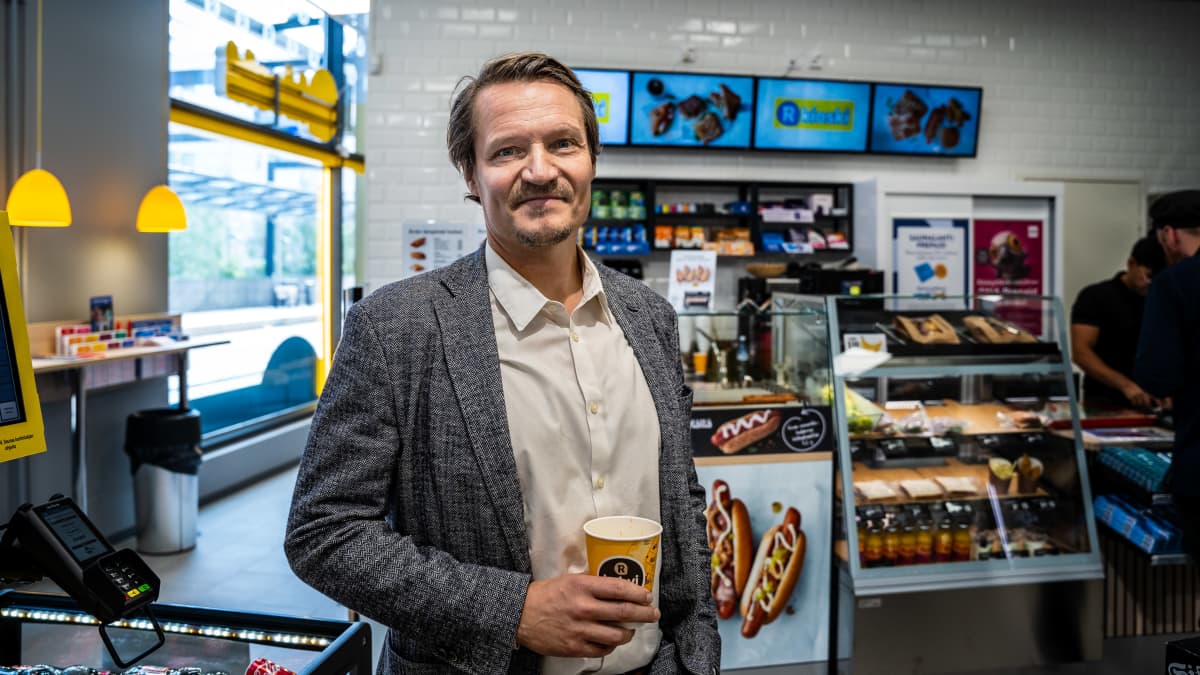 En vit medelålders man tittar mot kameran. Han håller i en kaffekopp och står inne i en R-kiosk. 