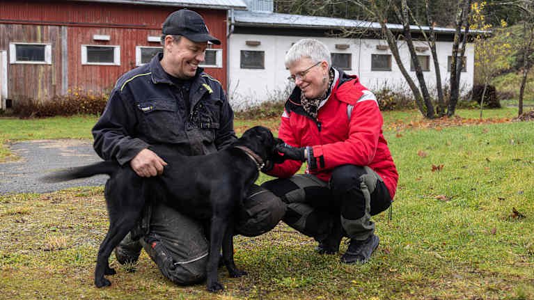En man och en kvinna står på huk med en svart hund på gården till ett lantbruk.
