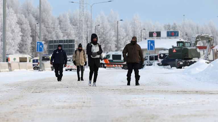 Rajavartija ja poliisi saattavat kahta tummaihoista miestä lumisella tiellä, joka johtaa Vartiuksen rajanylityspaikan Venäjän puoleiselle rakennuksille.