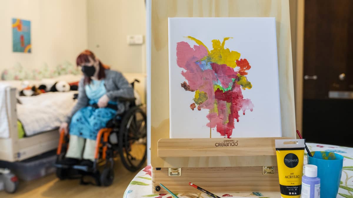 Etualalla kehitysvammaisen Eveliinan maalaama taulu, taustalla Eveliina pyörätuolissa.