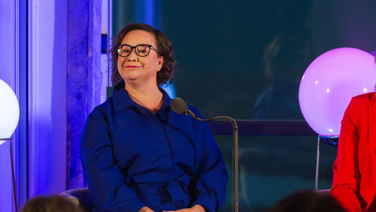 Sinisiin pukeutunut nainen istuu ja hymyilee leveästi, edessään mikrofoni.