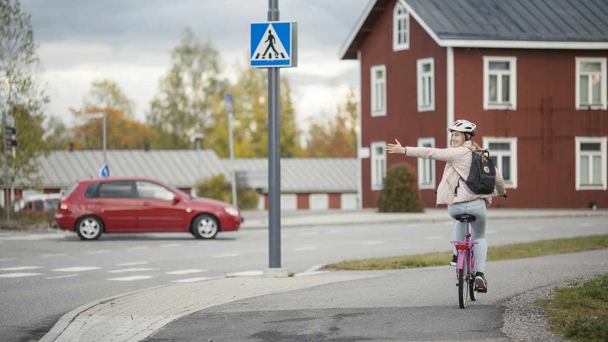 Risteyksessä auto ajaa oikealle ja pyöräilijä näyttää kädellä kääntymismerkin vasemmalle.  