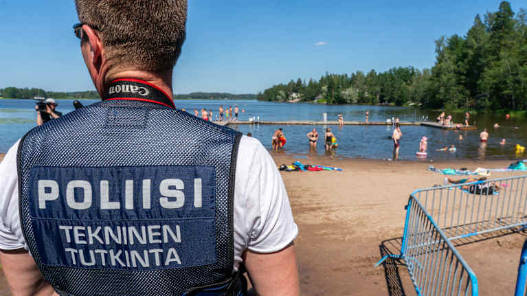 Poliisin tekninen tutkija uimarannalla, taustalla ihmisiä nauttimassa aurinkoisesta päivästä.