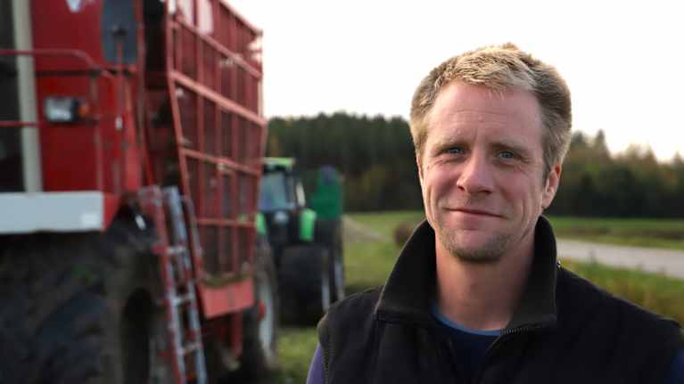 En man med brunljust hår tittar rakt in i kameran. Bakom sig har han en stor sockerbetsupptagare och en traktor med släp.