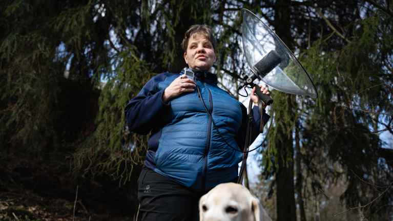 Nainen sinisessä takissa seisoo metsässä ja pitää kädessään parabolimikrofonia. Vieressä istuu vaalea labradorinnoutaja.