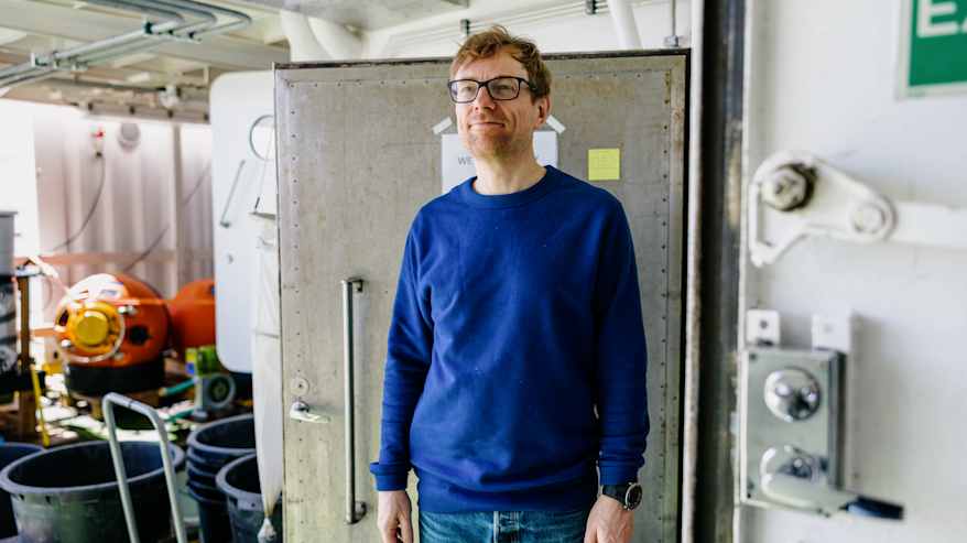 Kuvassa henkilö seisoo teollisessa ympäristössä, jossa taustalla näkyy metallisia ja muovisia elementtejä.