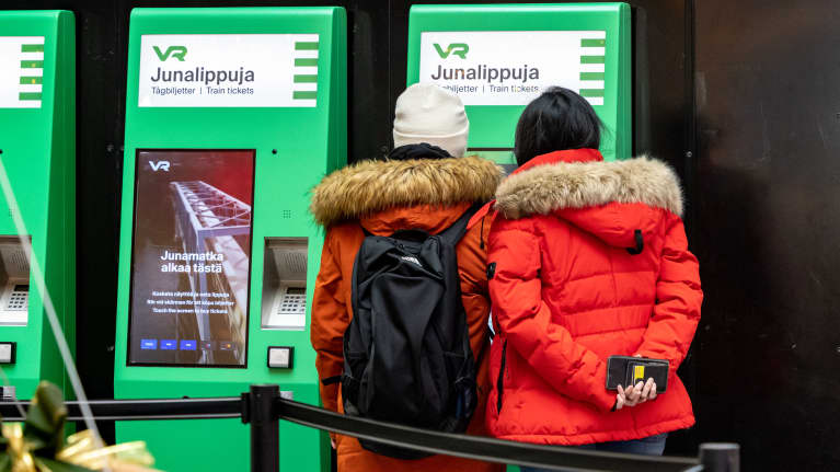 Kaksi henkilö ostamassa VR:n automaatilla junalippuja.