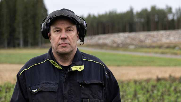 En man står klädd i arbetskläder och hörselskydd, med en bakgrund av skog och fält.
