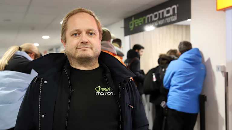 Mustaan paitaan ja talvitakkiin pukeutunut mies katsoo kameraan. Taustalla tiski, jonka ääressä paljon ihmisiä ja yläpuolella teksti Green Motion.