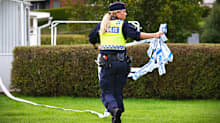 En polis går på en gräsmatta och håller i polistejp. Hon har ryggen mot kameran och tittar mot sidan så att hennes ansikte delvis syns.