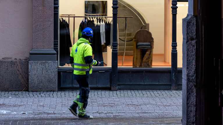 Rakennustyömies kävelee kadulla vaateliikkeen näyteikkunan ohi kaupunkimaisemassa.