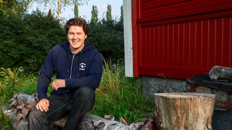 En person sitter på en stor trädstam intill en röd byggnad, med gräs och buskar i bakgrunden.