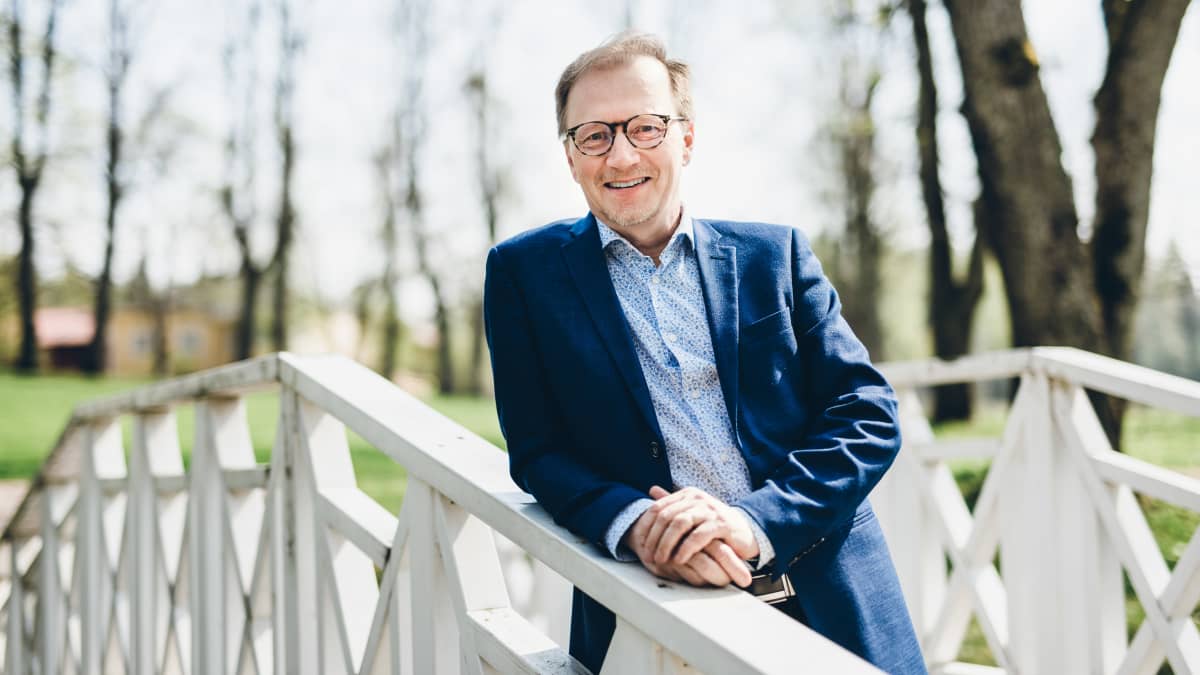 Keväisessä maisemassa pikkutakkiin pukeutunut mies hymyilee puisen sillan kaiteeseen nojaten.