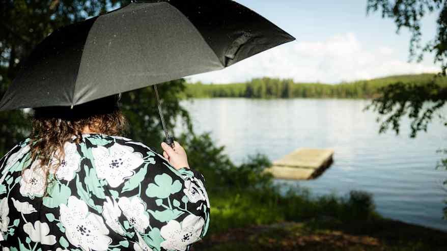 Kukkapaitaan pukeutunut nainen seisoo sateenvarjon kanssa ja katsoo järvelle. Sateenvarjo peittää naisen kasvot.