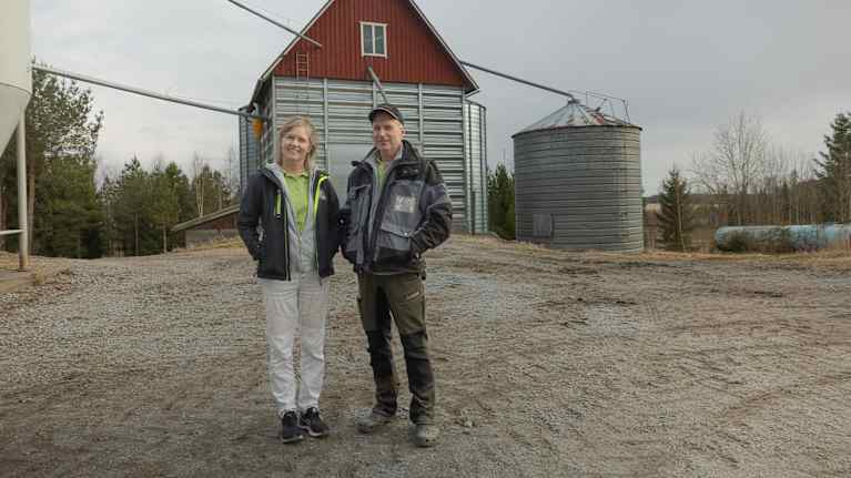 Två personer står framför en röd träbyggnad och en silo på en gårdsplan.
