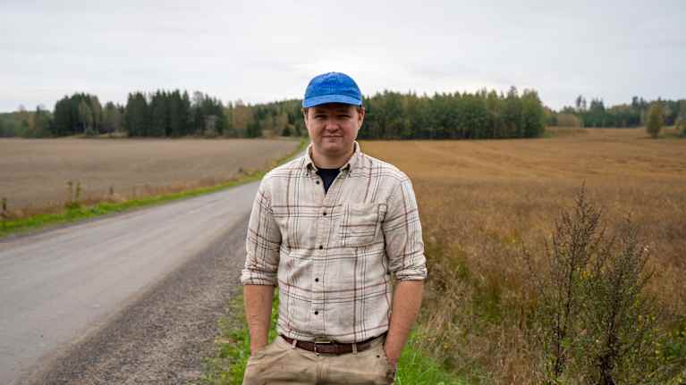 En ung man som ser mot kameran och har åkerfält och en sandväg i bakgrunden. 