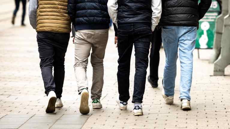 Four youths wearing down jackets, jeans and sneakers walking side-by-side on a city sidewalk.