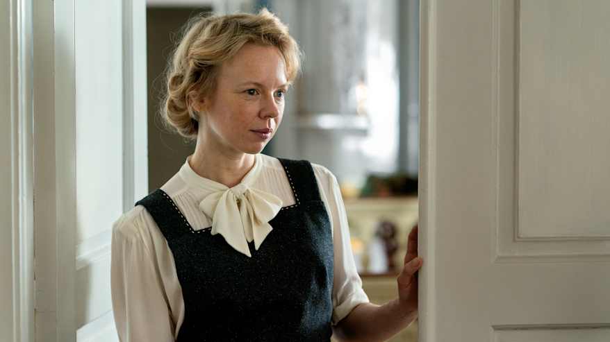 Henkilö levollinen ilme kasvoillaan ovenraossa 1940-luvun asussa, paitapusero jossa rusetti ja olkaimellinen hame, kuva Tove-elokuvan kuvauksista.